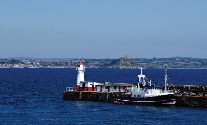 View to the north-east, from Newlyn Harbour to St. Michael's Mount, showing the lighthouse and Tidal Observatory to its right, both painted red and white. The white GPS antenna on top of the lighthouse platform can be clearly seen. (Source Wikimedia Commons.)